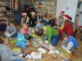 Dzieci ze szkoły podstawowej siedzą na podłodze i odwijają z papieru prezenty. Na ścianach wokół regały z książkami.