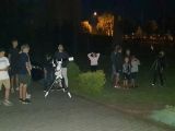 Grupa piętnastu osób stoj na trawniku i obserwuje nocne niebo. Pomiędzy nimi stoją trzy teleskopy.