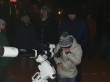 W nocy na podwórzu szkoły grupa osób obserwuje gwiazdy. Na pierwszym planie dziecko patrzy przez teleskop.