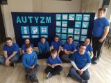 Grupa uczniów siedzących na podłodze w niebieskich koszulkach. W tle granatowa tablica