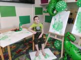 Chłopiec ubrany na zielono z odciska ślad stopy na białej kartce. W tle zielone plakaty i balony