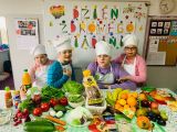 Dzieci siedzą przy stole, w tle napis Dzień Zdrowego Śniadania, na stole leżą owoce i warzywa. Uczniowie ubrani są w...