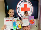 Dwie dziewczynki z dyplomami i kolorowymi obrazkami w dłoniach. W tle logo Polski Czerwony Krzyż