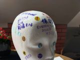Makieta głowy ze styropianu, ozdobiona i opisana przez uczniów kolorowymi pisakami. Na brystolu leży plansza pokazująca...