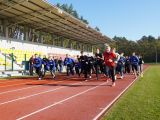 Na stadionie, na pomarańczowej bieżni zaczyna biec grupa ludzi w strojach sportowych (około 100 osób).