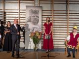 Mężczyzna w garniturze przemawia przez mikrofon. za nim banner z postacią króla z zarostem i w koronie. Obok uczennice w...
