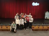 Grupa uczniów na scenie w teatrze. Za nimi bordowa kurtyna. Po bokach teatralne fotele