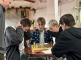 Kilkoro uczniów siedzi przy stoliku na szkolnej auli. Dwoje z nich gra w szachy, pozostali przyglądają się. Są skupeni i...