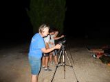 Kobieta w okularach robi zdjęcie nocnego nieba aparatem na statywie. Obok stoi dwóch chłopców przy drugim aparacie na...