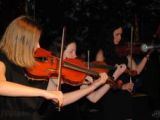 Trzy dziewczyny w czarnych sukienkach stoją w rzędzie na cenie i grają na skrzypcach.