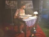 Przy stoliku na rynku siedzi kobieta w okularach i czyta książkę do mikrofonu. Na stoliku stoi zapalona lampka.