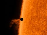 Z prawej strony, fragment Słońca, który zajmuje połowę zdjęcia. Z lewej strony przy Słońcu mały czarny punkt.