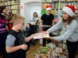 Uczeń szkoły podstawowej odbiera prezent od licealistki w czapce św. Mikołaja. W tle inne dzieci i dorośli.