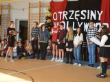Przy ścianie w rzędzie stoi siemioro licealistów. Pbok stoją dwie dziewczyny i trzymają mikrofon. Na ścianie napis...