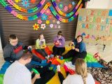 Dzieci bawią się siedząc na dużej kolorowej chuście. W tle kolorowe kwiaty i łańcuchy z papieru