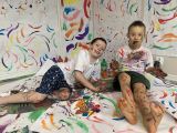 Ubrudzeni kolorowymi farbami chłopcy leżą na podłodze. W tle ściana z kolorowymi mazajami