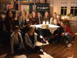 Dwanaście dziewczyn z liceum i dwie kobiety siedzą wokół stolika. Na stole stoją dwie zapalone świece.
