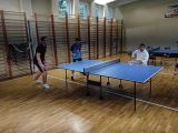 Dwuch licealistów w strojach sportowych stoi po jednej stronie stołu do tenisa. Pośrodku stołu sidzi chłopak w białej bluzie.