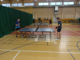 W ali gimnastycznej w rzędzie stoją trzy stoły do tenisa stołowego. Przy stołach uczniowie grają w ping-ponga.