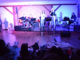 W sali na scenie dwie kobiety śpiewają do mikrofonu, trzech mężczyzn za nimi gra na gitarach. Przed sceną na materacach...
