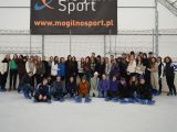 Duża grupa licealistów stoi przy barierce na lodowisku. Dziesięć osób siedzi na lodowisku. Wszyscy mają na nogach łyżwy.