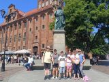 Grupa ludzi przed pomnikiem Mikołaja Kopernika, za nimi duży budynek z czerwonej cegły